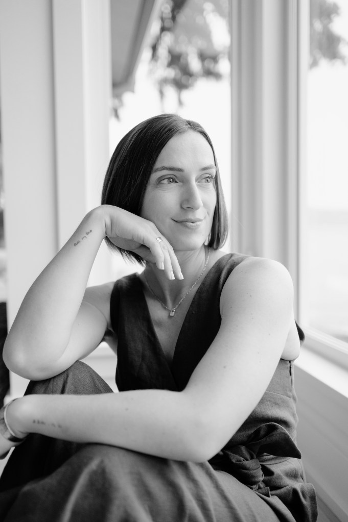 Kate v Eckland, a white woman with brown hair cut into a bob, sits in front of a window with one arm draped across her knees. The other arm is on a knee with her hand resting below her chin